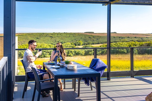 Couple laugh over drinks on the decking of their lodge at Gwel an Mor
