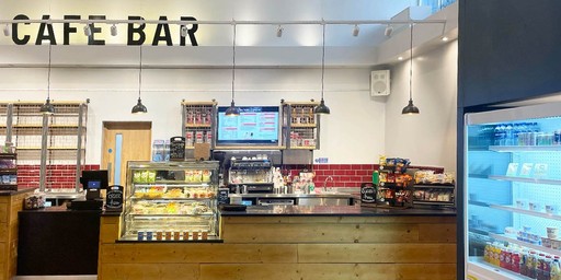 Café bar counter with food displays, drinks fridge, and menu screens on the wall behind.