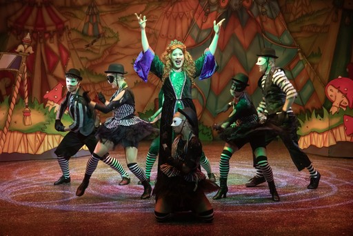 A woman in a colourful costume performs on stage with masked dancers in striped outfits.