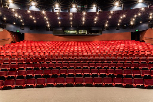 Empty rows of red and black theatre seats facing a stage in an auditorium.