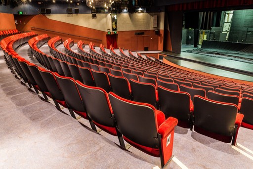 Empty rows of red and black theatre seats facing a stage in an auditorium.