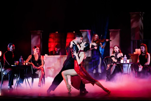 A couple passionately dances tango on stage, surrounded by musicians and onlookers in dim lighting.