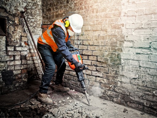 Construction worker using a heavy-duty breaker tool while wearing PPE and high-visibility clothing.