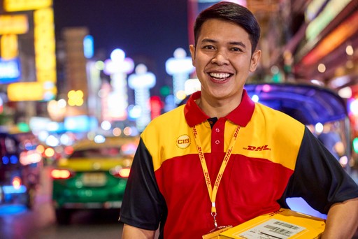 DHL Express courier wearing a branded red and yellow uniform is holding a parcel on a busy city street at night, surrounded by colourful lights and traffic.