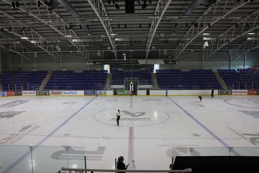 Empty indoor ice rink with two skaters practicing; rows of blue seats in the background.
