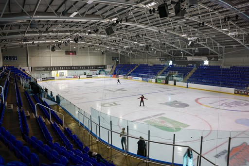 A few people skate on a large indoor ice rink with empty blue seats around it.