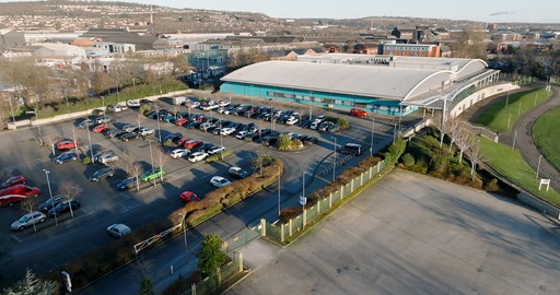 A large parking lot beside a modern building, with hills and houses in the background.