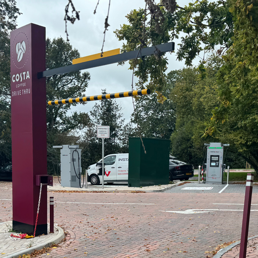 The Costa Coffee Drive Thru entrance in Bromsgrove features prominently beside two InstaVolt ultra-rapid chargers. A branded van and EV are parked at the charging bays, surrounded by trees and signage marking the drive thru route.