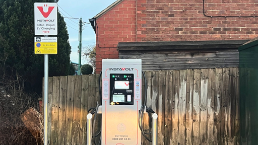 An InstaVolt ultra-rapid EV charging unit with dual connectors, located next to a brick building and wooden fence. A nearby sign indicates EV-only parking, CCTV operation, and 24/7 access.