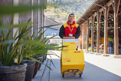 DHL Express courier transporting multiple yellow DHL Express boxes on a trolley in an outdoor setting with industrial-style buildings and greenery in the background.