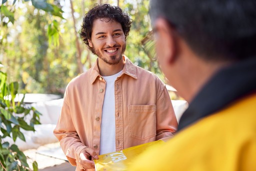 DHL Express courier delivering a yellow DHL Express Envelope 1 to a recipient wearing a light peach button-up shirt in an outdoor setting with greenery.