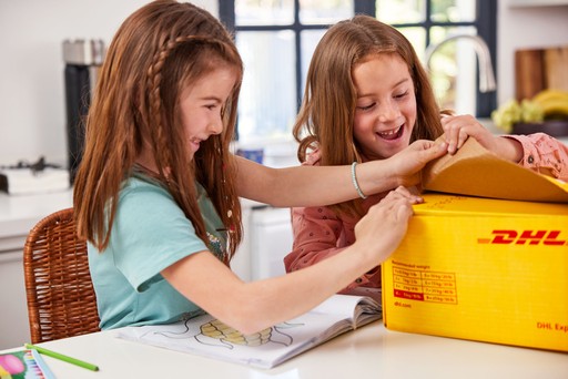 Two children opening a yellow DHL Express Box 3 on a table inside a home.