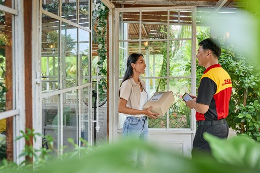 DHL Express courier completing a parcel delivery at a bright, plant-filled entrance while the recipient holds the package.