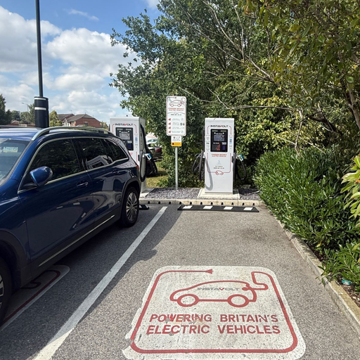 A blue electric vehicle is connected to InstaVolt’s dual rapid chargers in a marked EV bay. The setting features trees, signage, and convenient charger placement in a clean, organised car park.