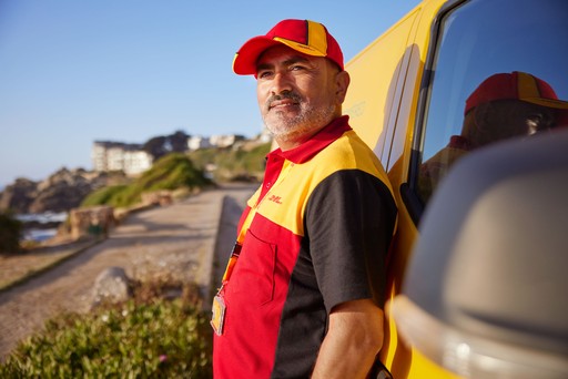DHL Express courier in a red and yellow uniform standing beside a yellow DHL delivery van on a sunny roadside.