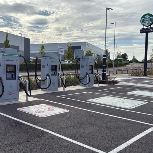 A row of InstaVolt ultra-rapid chargers located next to a Starbucks drive thru at Bell Farm Way, Kempston. Marked EV bays and a modern industrial estate are visible in the background.