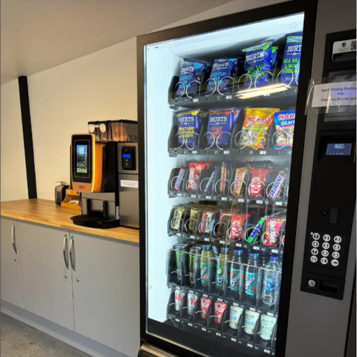 An indoor refreshment area featuring a fully stocked vending machine offering a range of crisps, sweets, chocolates, bottled drinks, and energy drinks. To the left, there is a bean-to-cup coffee machine positioned on a wooden countertop, offering freshly brewed coffee options. The space is clean and modern, providing convenient grab-and-go options for EV drivers.