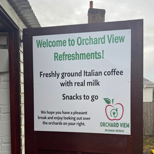 A sign at Orchard View Devon welcomes visitors with details about available refreshments. It advertises freshly ground Italian coffee made with real milk and a selection of snacks to go. The message encourages visitors to enjoy their break while overlooking the orchards. The Orchard View logo, featuring an apple with an electric plug, is visible at the bottom.