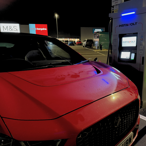 Nighttime scene of a red EV connected to an InstaVolt charger outside a retail park in Uckfield, with M&S and Home Bargains visible in the background.