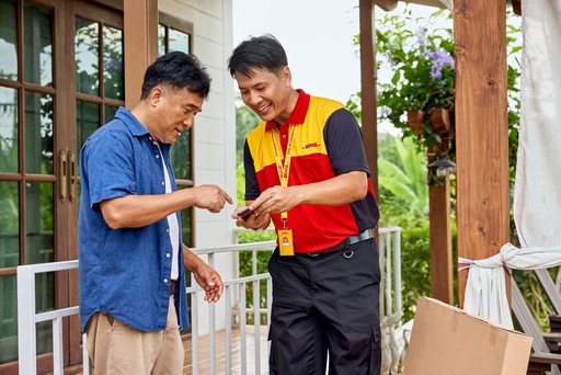 A DHL Express courier in a red and yellow uniform is assisting a customer on a residential porch, reviewing delivery information together.