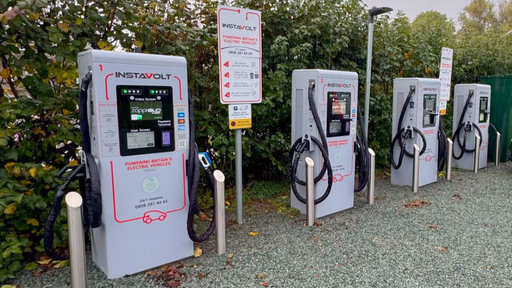 A row of four InstaVolt ultra-rapid EV chargers installed on a gravel forecourt, bordered by dense greenery under a grey, overcast sky. Located at The Pound Bed and Breakfast near the A438 in Whitney-on-Wye.