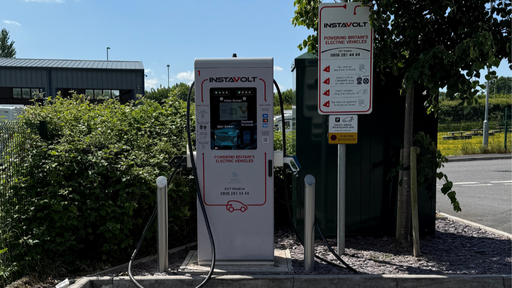 A wide view of an InstaVolt electric vehicle charger in Ilminster, installed beside a tree with visible instructions, cables, and clearly marked parking for EV use only. The background includes a business park and green hedging, giving it a practical yet scenic setting.