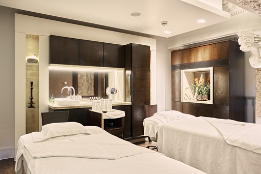 The image showcases a luxurious spa treatment room at the Sofitel. The room is bathed in soft, warm light, creating a serene and relaxing atmosphere. Two pristine white massage tables dominate the center of the room, each adorned with plush white linens and fluffy pillows, inviting guests to unwind and indulge in a pampering spa experience. A large, wooden cabinet with glass doors stands to the right, possibly housing various spa products or amenities. The room's overall design is sophisticated and elegant, with a focus on creating a tranquil and rejuvenating environment.