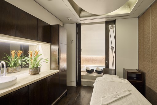 This image showcases a serene and inviting spa treatment room. The room is bathed in soft, natural light that streams in through a large window, overlooking a tranquil outdoor space. A comfortable massage table, draped in crisp white linens, takes center stage, inviting guests to relax and unwind. A sleek, modern vanity with a sink and mirror adds a touch of elegance to the space. Lush potted plants add a touch of greenery and freshness to the room, contributing to the overall sense of tranquility and well-being.