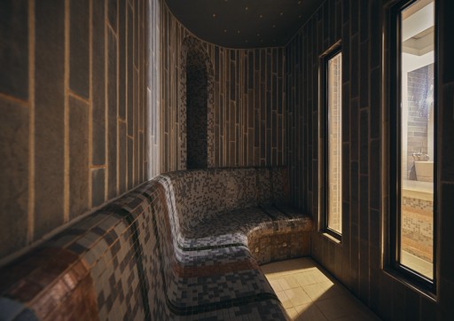 This image showcases a serene and dimly lit Turkish Hammam, also known as a Turkish bath. The room features a curved wooden bench lined with plush cushions, inviting guests to relax and unwind. The walls are adorned with intricate wooden panels, creating a warm and inviting atmosphere. A large window offers a glimpse of the outside world, allowing natural light to filter into the space. The overall ambience is peaceful and tranquil, perfect for a rejuvenating spa experience.