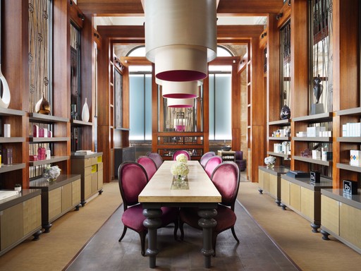 This image showcases the elegant waiting area of the Sofitel Spa. The room features a long, rectangular table with a white marble top, surrounded by a row of plush, velvet-covered chairs in a striking shade of pink. The walls are adorned with wooden paneling and shelves filled with various objects, creating a sense of sophistication and refinement. Large, cylindrical pendant lights hang from the ceiling, casting a warm and inviting glow over the space. The overall ambiance is serene and luxurious, setting the stage for a pampering spa experience.