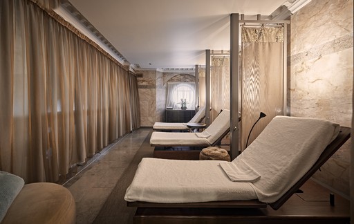 This image showcases a serene and inviting relaxation area within the Sofitel Spa. The room is bathed in soft, natural light, creating a calming atmosphere. A row of comfortable loungers, draped in plush white linens, invite guests to unwind and relax. The walls are adorned with warm, earthy tones and textured finishes, adding to the overall sense of tranquility. The overall design is minimalist and elegant, providing a peaceful escape from the hustle and bustle of everyday life.