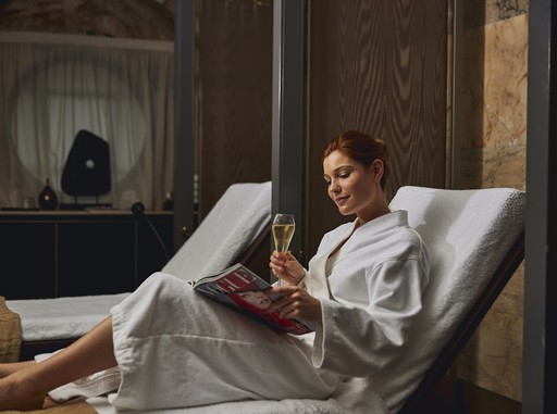 This image captures the essence of relaxation and indulgence at the Sofitel Spa. A woman, wrapped in a plush white robe, is seen lounging on a comfortable chair. She's enjoying a glass of champagne while engrossed in a magazine, creating a picture of pure tranquility. The soft lighting and muted color palette contribute to the serene atmosphere of the spa.
