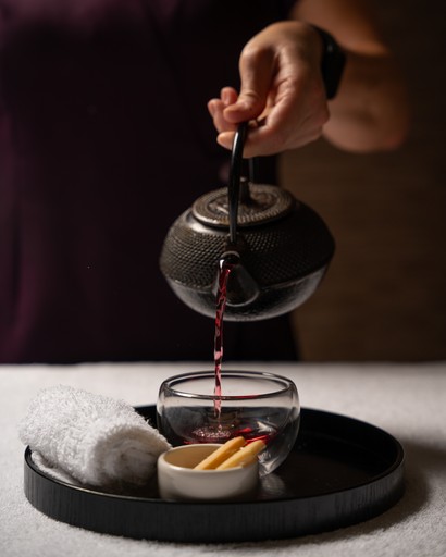 This image showcases the ritual of preparing a soothing herbal tea at the Sofitel Spa. A therapist's hand is pouring steaming tea from a traditional cast-iron teapot into a delicate glass cup. The tea is accompanied by a small bowl of snacks, likely to be enjoyed after the treatment. The overall setting, with its soft lighting and minimalist aesthetic, creates a sense of tranquility and well-being.