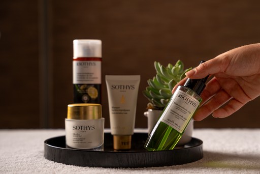 This image showcases a selection of luxurious skincare products used at the Sofitel Spa. A hand is reaching for a bottle of green serum, highlighting the range of treatments and products available. The products are displayed on a tray, alongside a small potted plant, creating a visually appealing and inviting presentation.