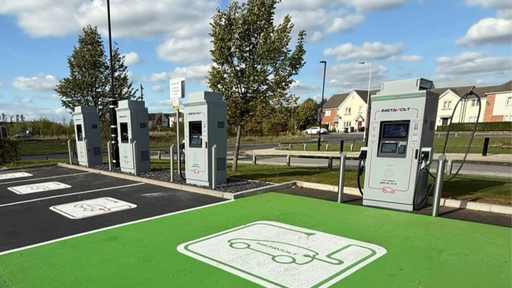 Wide-angle view of multiple InstaVolt ultra-rapid chargers set beside green and black EV bays, with nearby houses and trees under a clear blue sky.