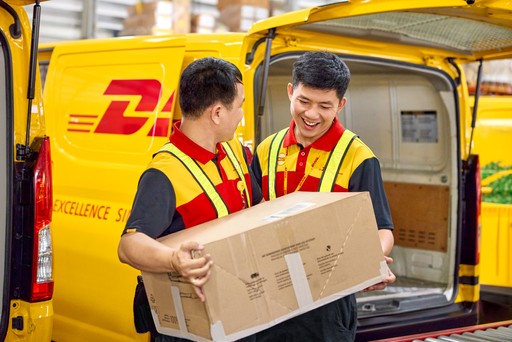 Two DHL Express couriers are handling a large cardboard box during a parcel loading process beside a yellow DHL Express delivery van.
