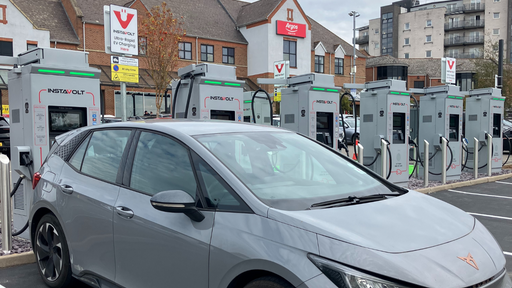 A grey electric car is charging at InstaVolt ultra-rapid chargers outside a shopping area featuring Argos, with several charging units available on-site in Northampton.