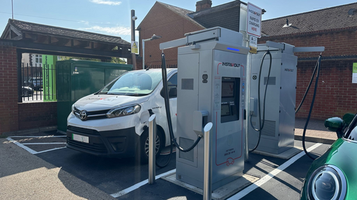 This image shows a pair of InstaVolt chargers actively in use, positioned near a brick building and a Co-operative Food store. A branded Toyota van and a green EV are parked and charging.