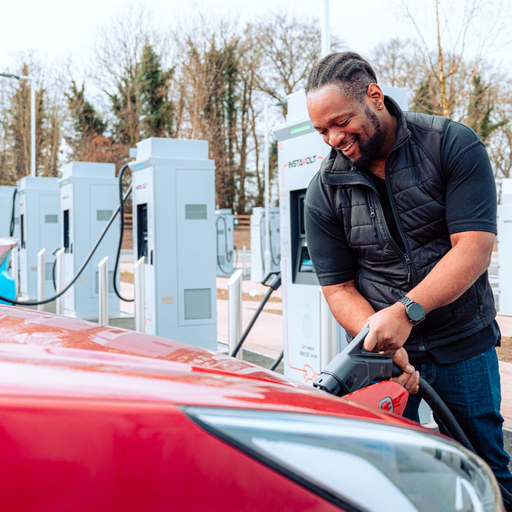 A man smiling while plugging his electric vehicle into an InstaVolt ultra rapid charger, with modern charge points in the background and a tree-lined setting.