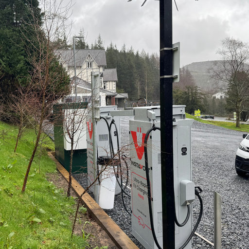 Angled view of the InstaVolt charging station showing dual ultra-rapid chargers, cable management arms and the Plas Weunydd Hotel nearby.