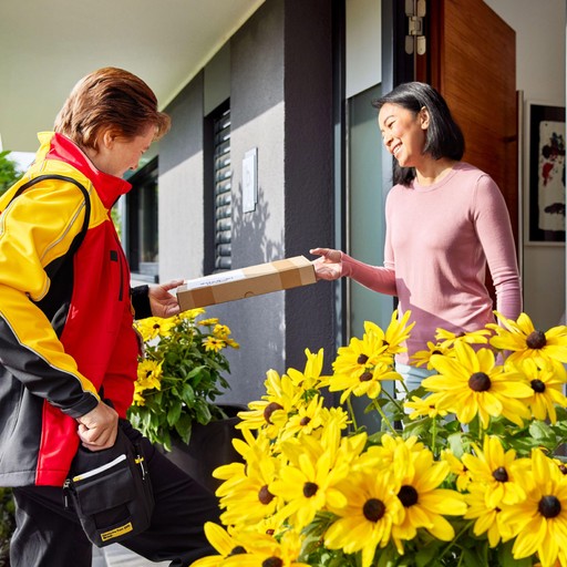 DHL Express courier delivering a parcel to a customer at a residential doorstep surrounded by bright yellow flowers.
