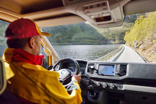 DHL Express courier driving a yellow delivery van along a scenic route with a view of water and green hills through the windshield.