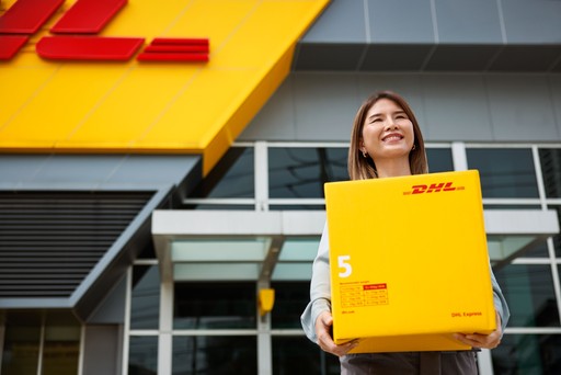 Person holding a large yellow DHL Express Box 5 in front of a DHL Express Service Centre with the iconic yellow and red branding.