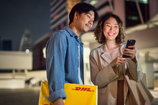 Two individuals standing outdoors at night, one holding a yellow DHL Express Envelope 1 while the other looks at a smartphone.