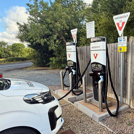 A white EV charges at one of the InstaVolt rapid charging units at Alcester Road Service Station. The location is positioned beside the A422, with rural scenery and trees in the background, offering easy roadside access.