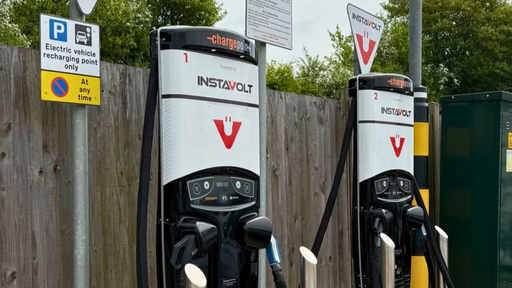 A pair of InstaVolt rapid electric vehicle chargers located at Alcester Road Service Station. Signage includes usage instructions and dedicated EV parking. The chargers are backed by a wooden fence and surrounded by greenery.
