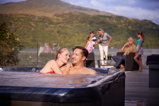 Couple sitting in Hot Tub while their family relax on the veranda of their lodge at Loch Lomond.