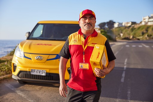 DHL Express courier in a red and yellow uniform carrying a small yellow DHL Express Box 2 while standing in front of a yellow DHL delivery van on a sunny roadside.
