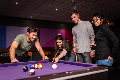 Four young adults, two male, two female, playing a game of pool with one of the girls taking her go while the others watch