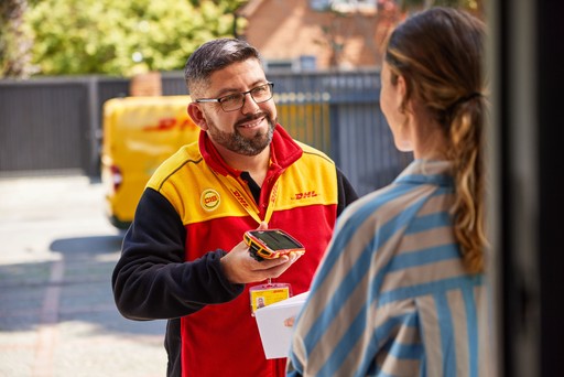 DHL Express courier assisting a customer with parcel delivery and electronic signature outside a residential property, with a yellow DHL van visible in the background.